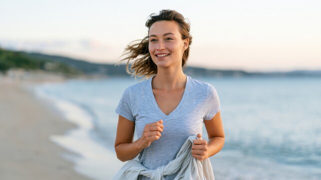 Young woman with long hair wearing a gray t-shirt walks along a sandy beach during sunset, ocean waves gently lapping at the shore in the background