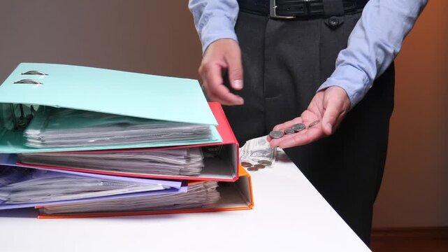 Businessperson by a stack of ring binders counts coins with banknotes and loose change on the table, conveying budgeting, financial strain, bookkeeping and cash flow concerns
