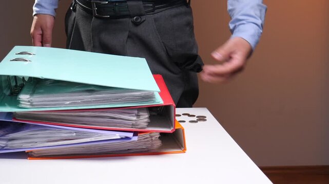 Employee pulling out empty pocket with a few coins, standing next to a stack of ring binders, depicting financial crisis, poverty, and low wages in business