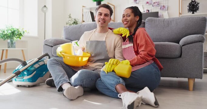Couple cleaning home with housekeeping vacuum near sofa. Smiling man and woman in aprons and gloves sit on floor with bucket and spray bottle after chores in a bright apartment.