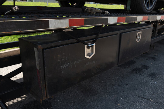 Close-up of a black metal toolbox mounted under a flatbed trailer, secured with a metal latch, featuring red and white reflective safety striping, highlighting industrial transport safety