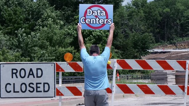 Man holding No Data Centers sign construction site barricades protesting AI tech infrastructure power grid development environmental impact zoning