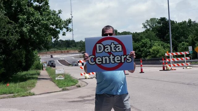 Man holding No Data Centers sign construction site barricades protesting AI tech infrastructure power grid development environmental impact zoning