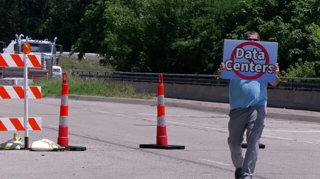 Man holding No Data Centers sign construction site barricades protesting AI tech infrastructure power grid development environmental impact zoning