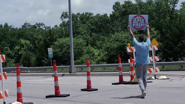 Man holding No Data Centers sign construction site barricades protesting AI tech infrastructure power grid development environmental impact zoning