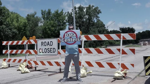 Man holding No Data Centers sign construction site barricades protesting AI tech infrastructure power grid development environmental impact zoning