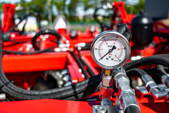 Detailed close-up view of a round pressure gauge mounted on a metal hydraulic manifold, surrounded by black hydraulic hoses and bright red painted machinery
