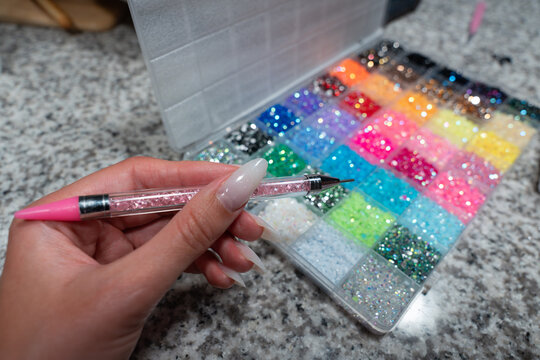 Hand using a rhinestone applicator tool with colorful crystals in the background. Popular bedazzling trend for customizing accessories, clothing, phone cases, and fashion item