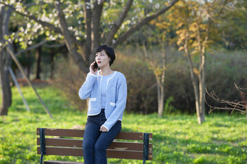 Fototapeta premium Young woman sits park bench, talking on phone. Sunlight filters through trees behind her. Her gaze is distant, thoughtful, focused. She wears casual knit top and jeans. Ideal for lifestyle, relaxation