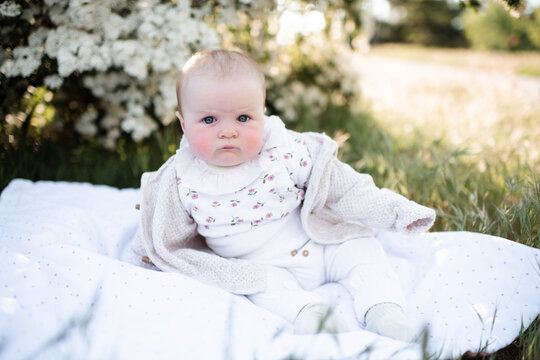 Funny baby girl under 1 year old with frowning face sitting on green grass lawn in park outdoors. Looking at camera. Childhood.