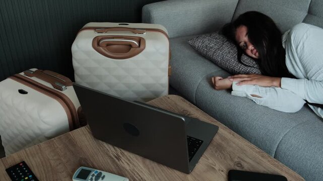 Digital nomad burnout and remote work fatigue. Woman lying on sofa near laptop and luggage feeling emotionally exhausted by constant travel