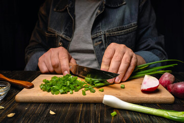 Hands are shown chopping green onions on a cutting board. Nearby, there are radishes and additional herbs. The setting is dark with a focus on the cutting task © Віталій Б.