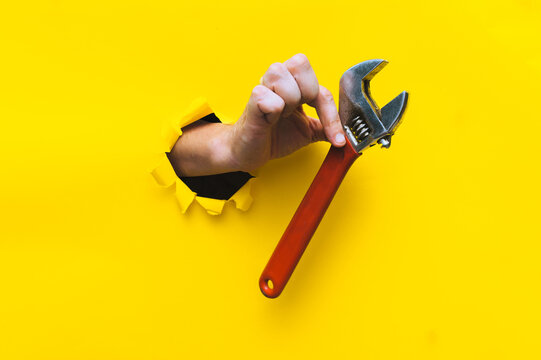 A right hand in a black knitted glove holds a red monkey wrench. Torn hole in yellow paper. The concept of a worker, labor migrant, a master of his craft. Copy space.