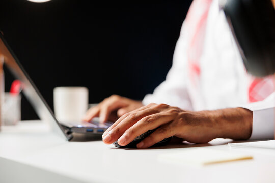 Close-up view of a pair of male hands using a wireless mouse and laptop, doing online research. Detailed shot of a person with Arabian clothing making use of the minicomputer on the table.