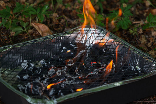 Burning charcoal glows brightly in a disposable grill over leaves and dirt.