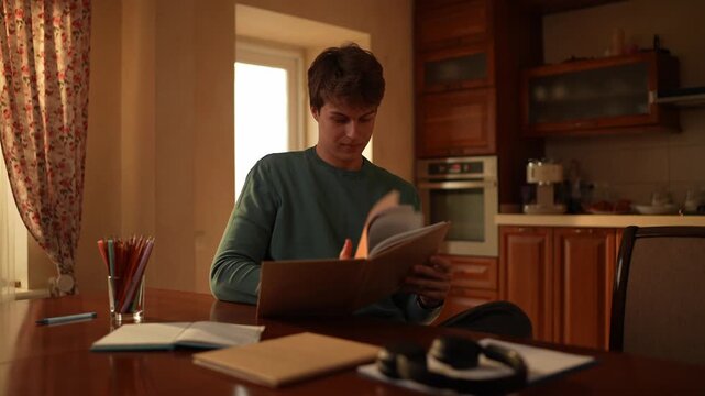 A young man sits at a kitchen table, looking through a notebook while surrounded by study materials and a window view