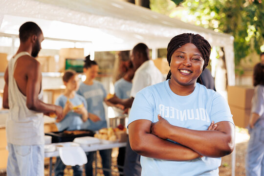 Enthusiastic black woman wearing blue t-shirt with arms crossed looks at camera. Portrait shot of african american female volunteer, ready to provide humanitarian aid to poor, needy and less fortunate