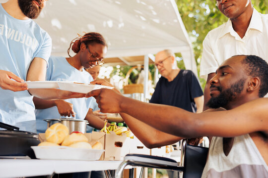 Helpful guy volunteer aids and provides free food to a poor needy african american man in wheelchair. Volunteers at an outdoor food bank provide hunger assistance to the crippled and underprivileged.