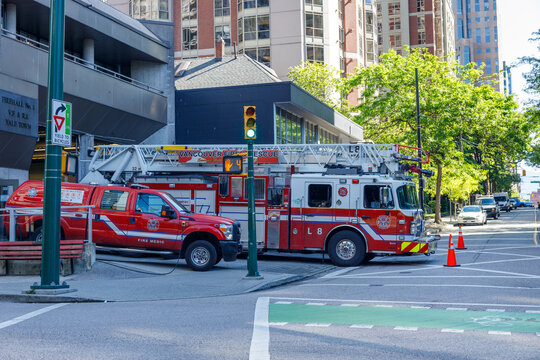 A Vancouver Fire Rescue ladder truck and a red fire medic pickup parked outside Firehall No. 1 in Yaletown, beside a downtown street and residential towers.