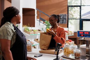 Obraz premium African American woman vendor at a small grocery store with a female customer, promoting healthy and sustainable produce. Young client with brown paper bag, unloading fresh lemons at counter.