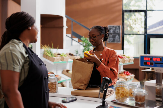 African American woman vendor at a small grocery store with a female customer, promoting healthy and sustainable produce. Young client with brown paper bag, unloading fresh lemons at counter.
