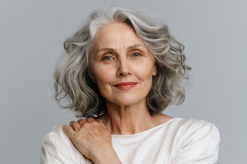Fototapeta premium Confident senior woman with natural gray wavy hair smiling softly in a studio portrait against a light neutral background. 