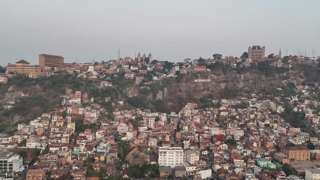 Cinematic aerial footage of the densely populated hillside city of Antananarivo, Madagascar, featuring historic palaces on the ridge during a hazy dusk or morning.