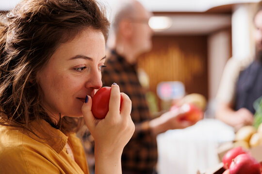 Woman smelling organic handpicked apples in zero waste supermarket while vendor talks with elderly customer in blurry background, client testing to see if local neighborhood shop fruits are fresh.