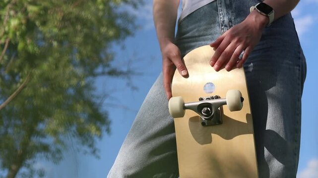 Young girl in white t-shirt and jeans holding skateboard against blue sky and trees. Bottom view. Stylish skateboarder girl posing with her board outdoors on a sunny day. Active leisure concept