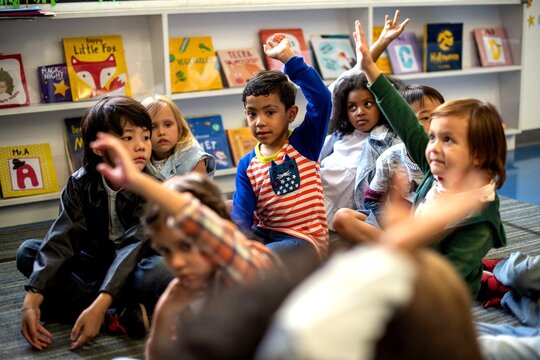 Diverse group of children sitting on the floor, raising hands in class.
