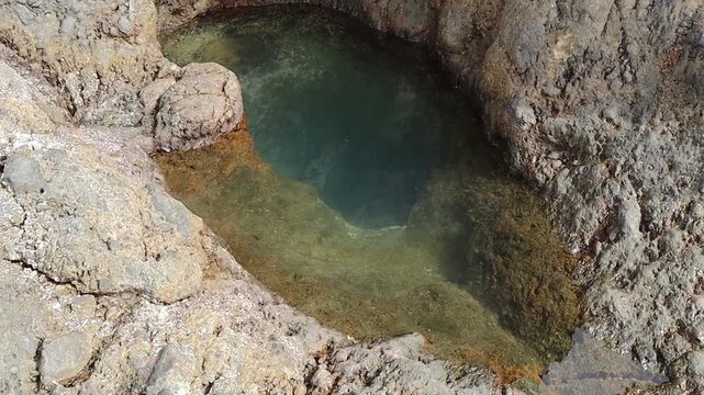 A small, shallow natural crevice in the rock filled with clear Atlantic Ocean water, with small fish swimming in it, on the island of Tenerife on a clear autumn day, viewed from above, close-up, in sl