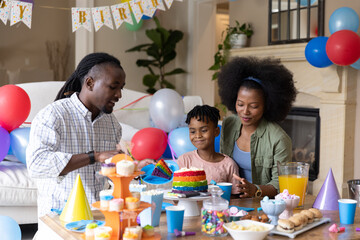 Obraz premium African American family celebrating birthday in living room cutting rainbow cake near cupcakes
