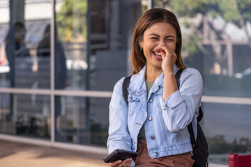 African American woman sitting on low ledge laughing, holding phone in denim jacket by glass facade © wavebreak3