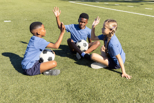 Diverse children sitting on grassy soccer field holding two black white soccer balls in blue shirts