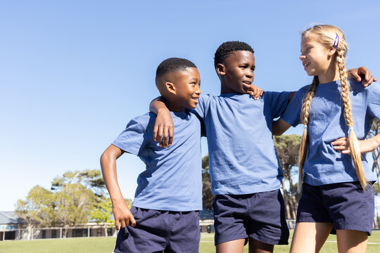 Diverse school children linking arms chatting on school field wearing blue tees and navy shorts