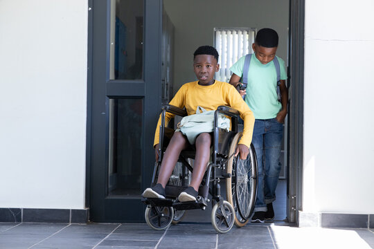 African American school-age boys pushing manual wheelchair through dark door at entrance, backpacks