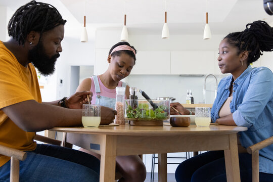 African family with teen daughter sharing salad from glass bowl with tongs at kitchen table