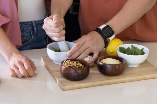 Couple crushing greens with pestle in ceramic mortar on board at kitchen counter pine nuts, cheese