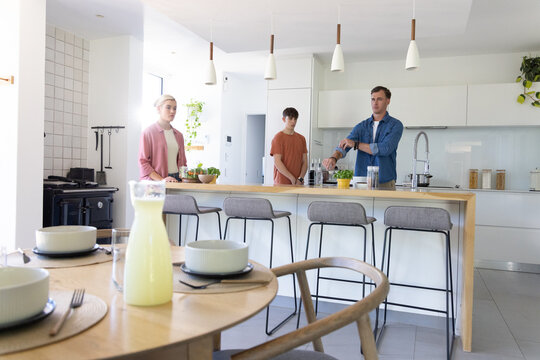 Family with mother in pink cardigan helping teenage son at kitchen island with coffee maker