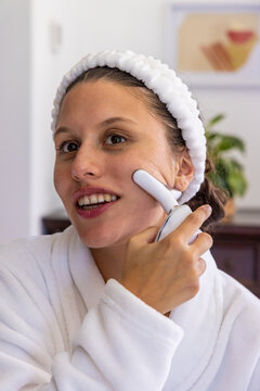 African American woman wearing bathrobe and headband applying skincare device to cheek at vanity