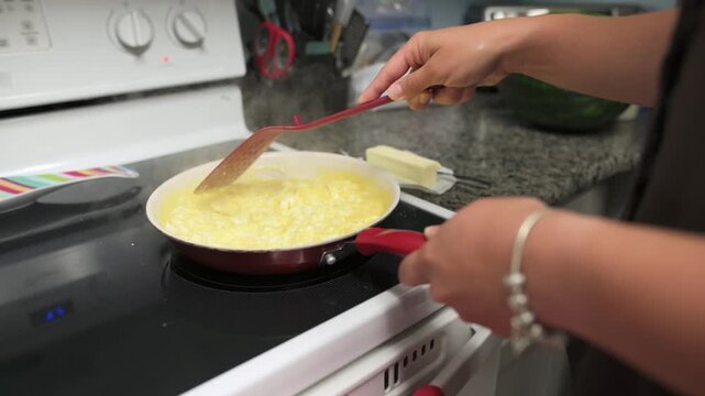 Woman prepares scrambled eggs in a red frying pan on a stovetop, using a spatula to stir the mixture, with butter and a watermelon placed on the countertop in a well-lit kitchen