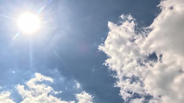 Bright white cumulus clouds fill a deep blue sky with a glowing sun shining intensely at upper right. Clouds slowly shift and expand across the frame in natural daylight.