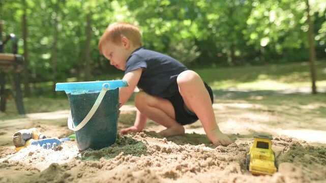 Young boy with red hair crouches in a sandbox, scooping sand with a green shovel into a blue bucket, surrounded by toy vehicles in a bright, sunny park environment