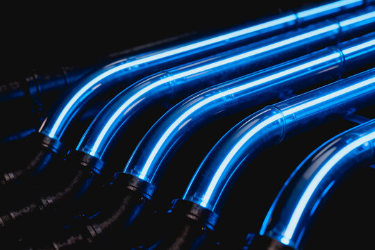 Parallel curved transparent pipes glowing bright blue against a dark background, forming an industrial technical structure.