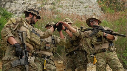 Fototapeta premium A diverse team of soldiers in full combat gear stacks hands together, demonstrating unity and strategizing for a military operation in a rugged environment.