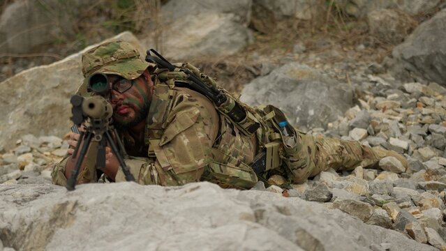 A special forces sniper with face paint lies prone on a rocky outcrop, providing overwatch and conducting reconnaissance during a covert military operation.