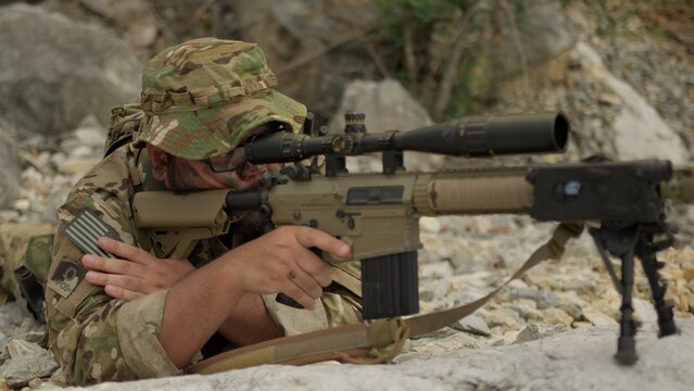 A military sniper in multicam camouflage aims his rifle from a concealed rocky position, conducting surveillance during a special operations mission.