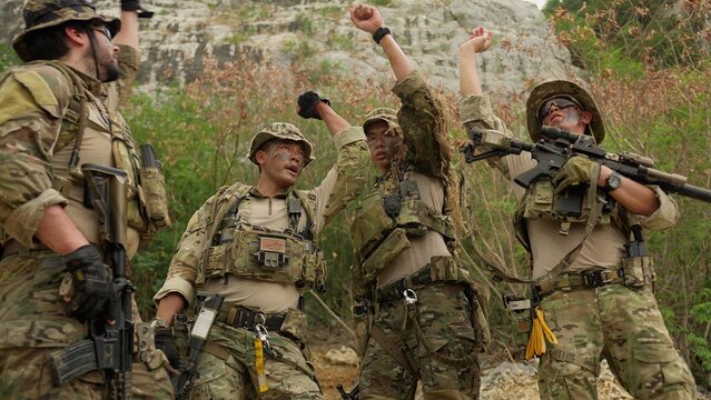 A diverse team of soldiers in full combat gear stacks hands together, demonstrating unity and strategizing for a military operation in a rugged environment.