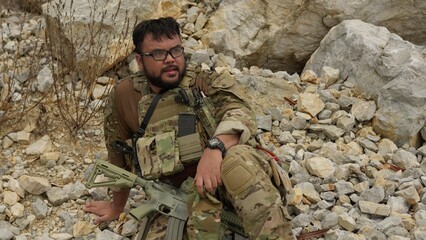 Obraz premium A man in tactical gear takes a break from a military simulation, resting with his rifle among the rocks of a rugged, outdoor training environment.
