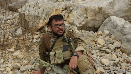 Obraz premium A man in tactical gear takes a break from a military simulation, resting with his rifle among the rocks of a rugged, outdoor training environment.